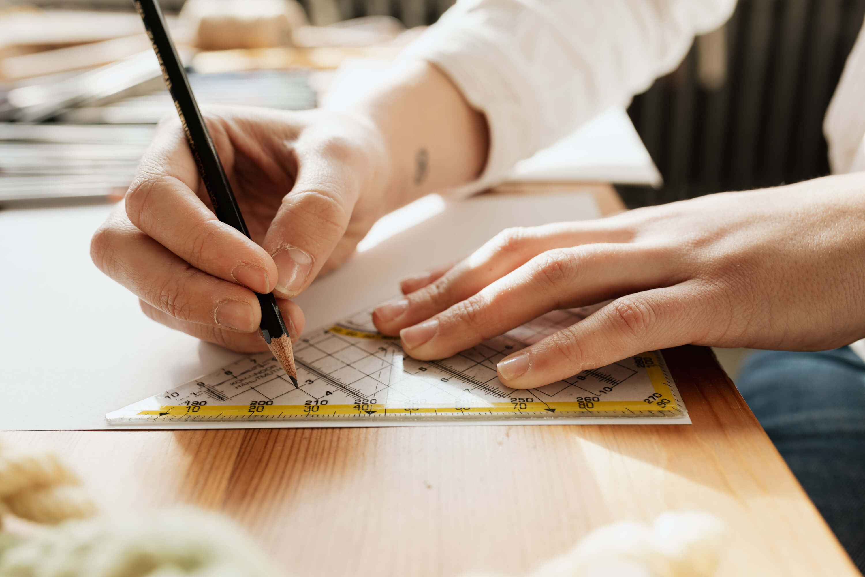 A pair of hands using a pencil and a set square on a white sheet of paper.